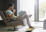 Ein junger Mann sitzt in einem Sessel im Wohnzimmer, liesst ein Buch und trinkt ein Getraenk.