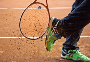 Nahaufnahme der Füße eines Spielers und eines Tennisschlägers. Durch Bewegung wird Schotter in die Luft gewirbelt.