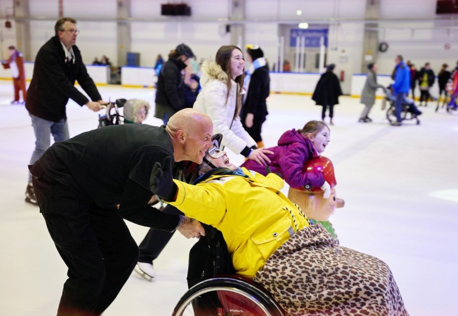 Eine Person im Rollstuhl und ihr Begleiter auf Schlittschuhen haben gemeinsam Spaß auf der Eisfläche.