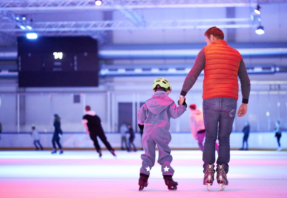 Ein Vater fährt mit seinem Kind an der Hand auf Schlittschuhen auf der Eisfläche der Eissporthalle. © Stiftung Eissporthalle Düsseldorf-Benrath