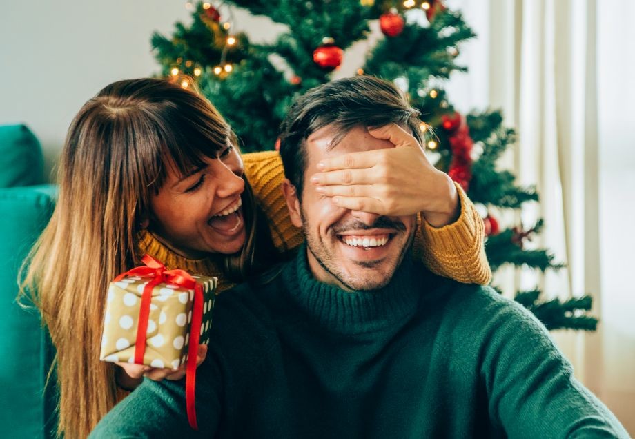 Schrottwichteln: Lachende Frau hält Mann die Augen zu und überreicht ihm ein Geschenk.