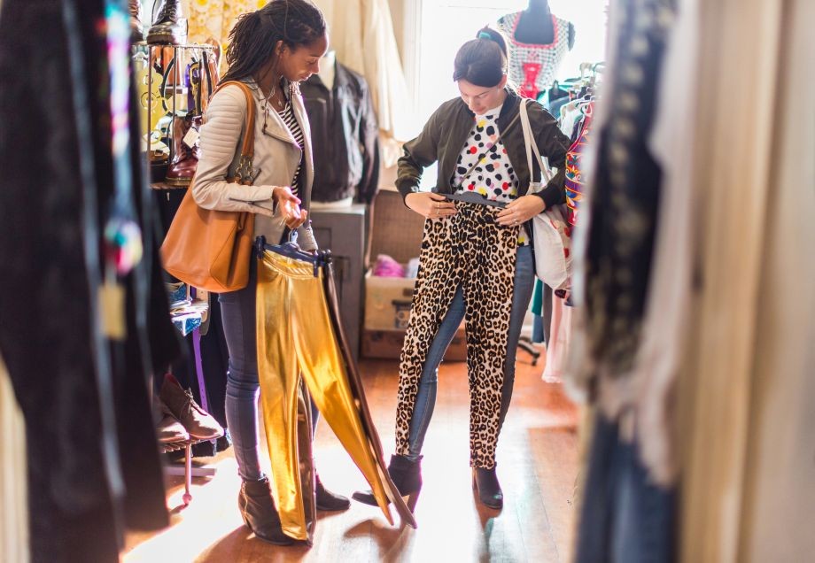 Zwei junge Frauen stöbern in einem Vintage-Laden und halten Second Hand-Kleidung in der Hand.