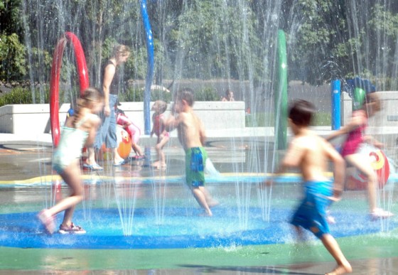 Kinder laufen auf einem Wasserspielplatz durch ein Fontänenfeld. 