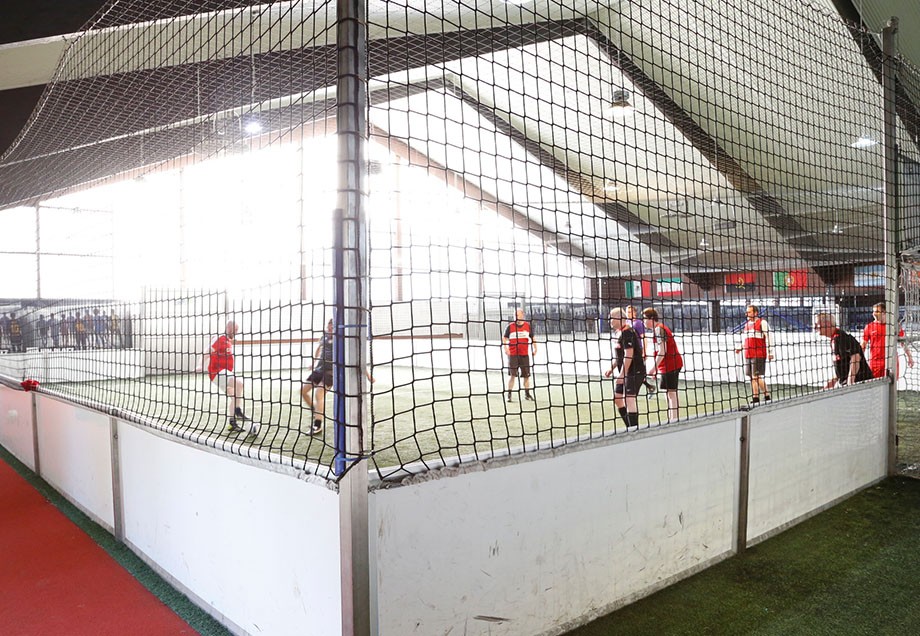 Indoorfußball in einem Feld, welches mit einem Netz eingegrenzt ist. In dem Feld spielen Menschen in roten und schwarzen Trikots Fußball.