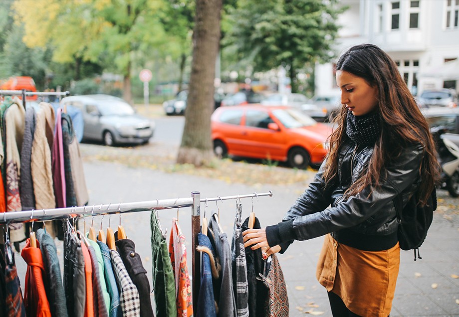 Eine Frau schaut sich Kleidung auf Kleiderstangen an, die im Rahmen eines Hofflohmarktes auf dem Bürgersteig ausgestellt sind.