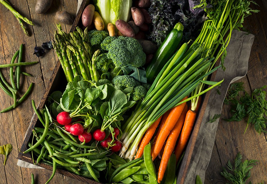 Blick von oben in eine Holzkiste mit Gemüse, unter anderem Brokkoli, Radieschen, Spargel, Möhren und Kartoffeln.