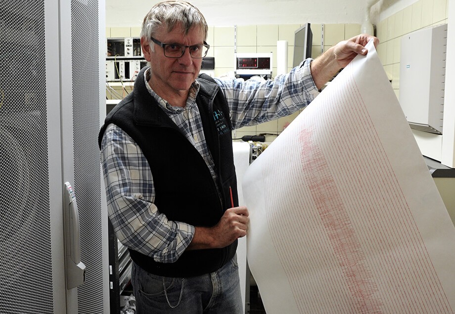 Ein in die Kamera schauender, bebrillter Mann hält einen großen Papierbogen, auf dem seismografische Aufzeichnungen festgehalten sind.