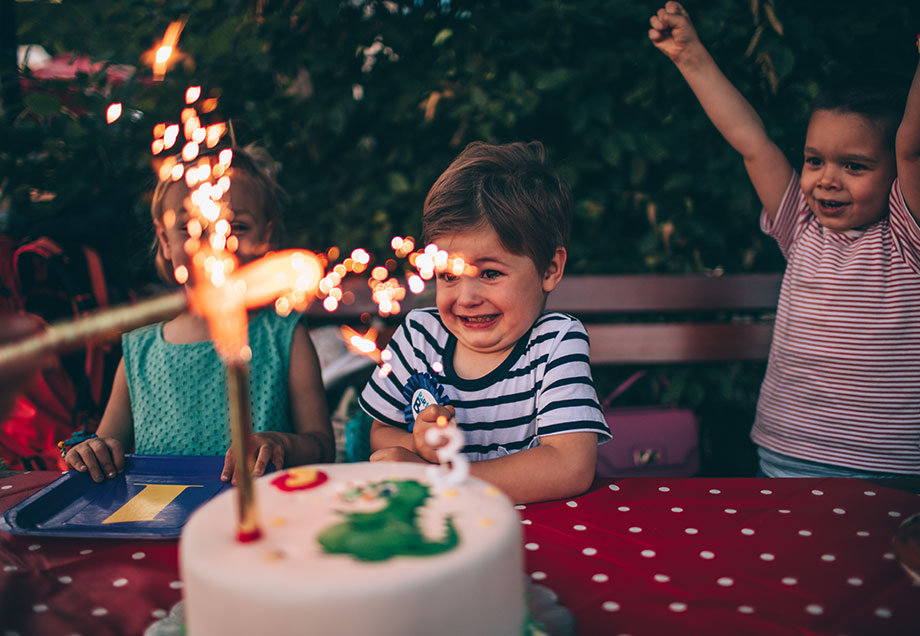 Kinder freuen sich über Funken sprühendes Feuerwerk auf weißem Geburtstagskuchen mit Drachen aus Zuckerguss. 