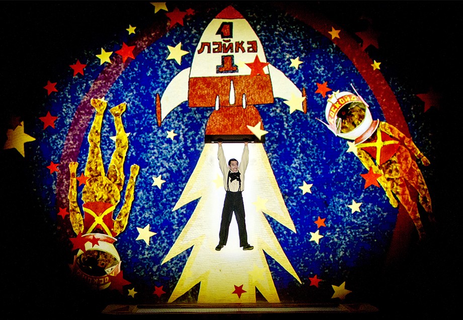 Bühnenszene: Im Hintergrund die Grafik einer Rakete mit russischer Beschriftung (Lajka) und zwei Astronauten. Ein realer Mensch hängt vermeintlich an der gezeichneten Rakete.