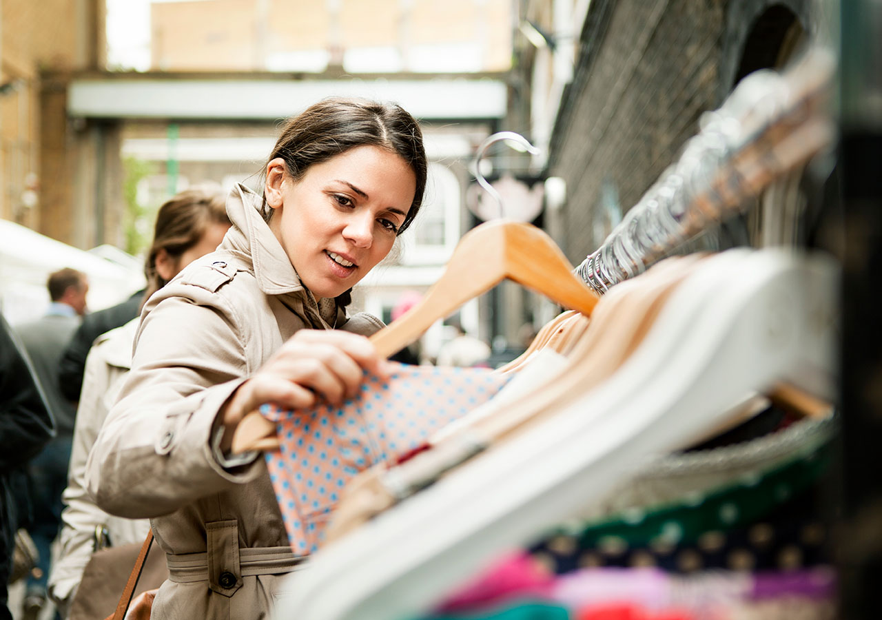 Eine Frau im beigefarbenen Trenchcoat betrachtet musternd eine lachsfarbene Bluse auf einem Bügel, die sie offenbar von einer vollgehangenen Kleiderstange auf einem Trödelmarkt genommen hat.
