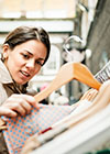 Eine Frau im beigefarbenen Trenchcoat betrachtet musternd eine lachsfarbene Bluse auf einem Bügel, die sie offenbar von einer vollgehangenen Kleiderstange auf einem Trödelmarkt genommen hat. 