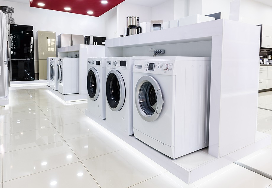 In einem Küchenfachgeschäft sind Waschmaschinen oder Trockner zum Verkauf in der Mitte des Verkaufsraums aufgestellt, umgeben von Kühlschränken und Küchenmöbeln