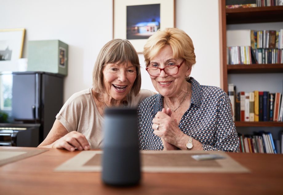 Zwei ältere Frauen sitzen begeistert vor einem Smart Speaker und entdecken dessen Funktionen.