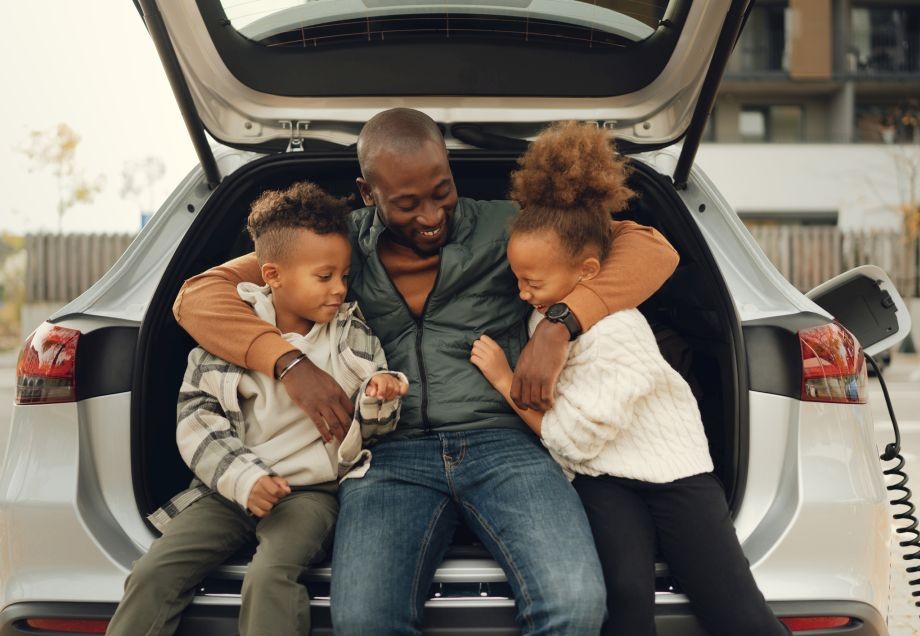 Ein Vater sitzt mit seinen zwei Kindern am Rand des offenen Kofferraums eines Elektroautos, während es lädt.