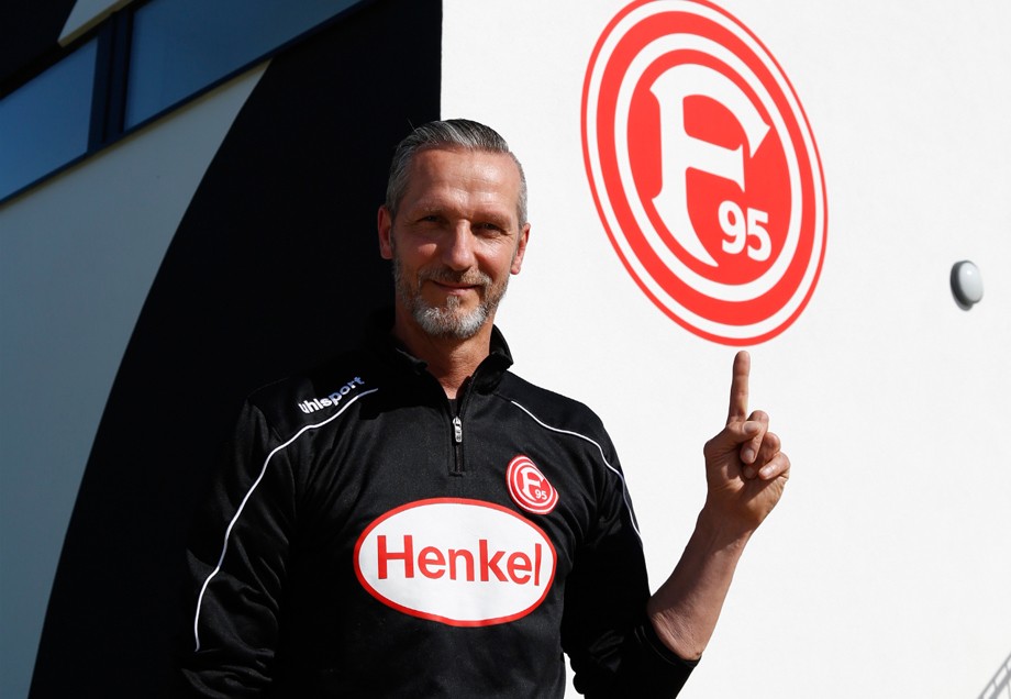 Carsten Fiedler zeigt auf das große Vereinslogo von Fortuna Düsseldorf an einer Wand des Nachwuchsleistungszentrums.