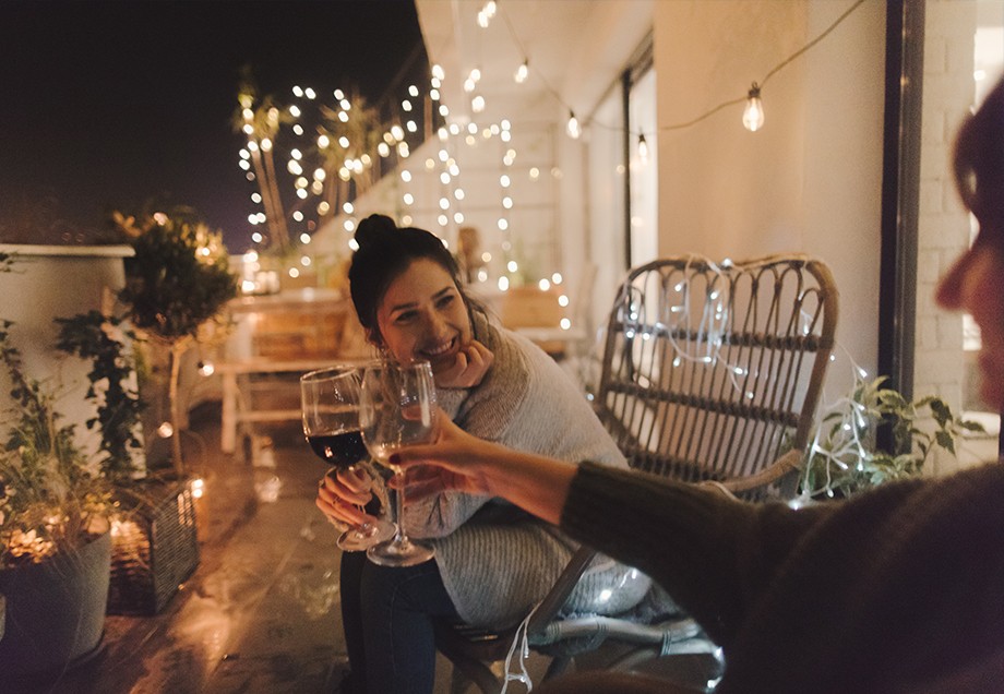 Freundinnen stoßen auf der mit vielen kleinen Lichtern dekorierten Terrasse mit einem Glas Rotwein an.