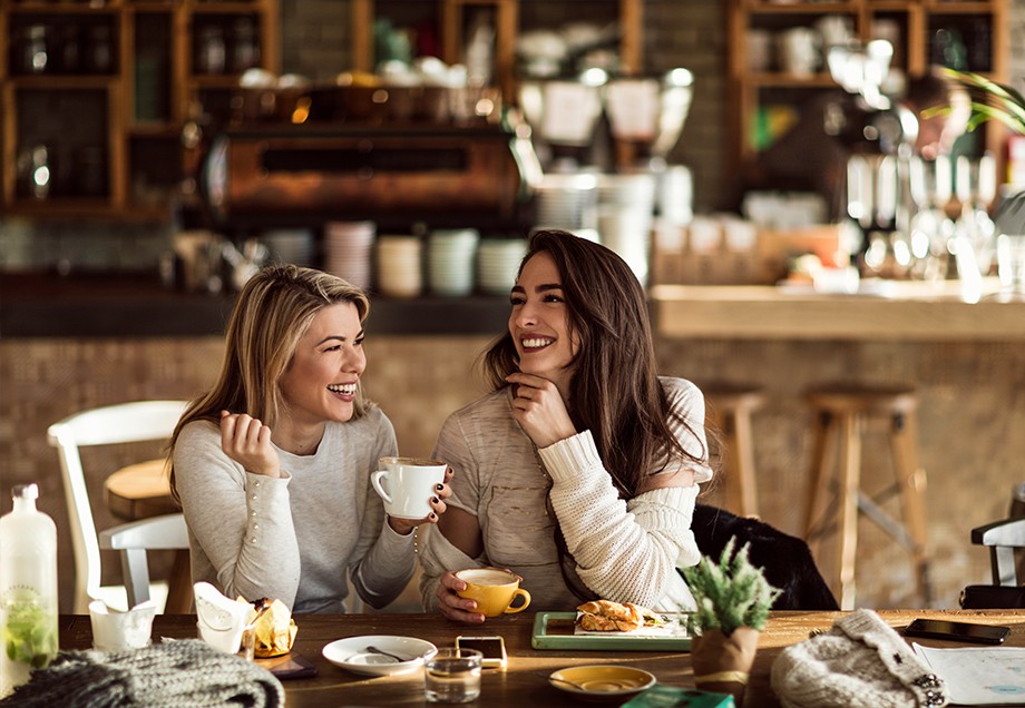 Zwei Freundinnen trinken lachend Kaffee in einem liebevoll dekorierten Café, in das warmes Sonnenlicht fällt.