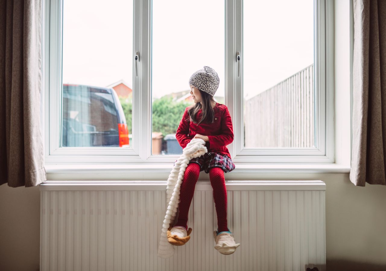 Ein Mädchen sitzt auf einer Fensterbank und wärmt sich an einer Heizung auf. © Tang Ming Tung / DigitalVision via Getty Images
