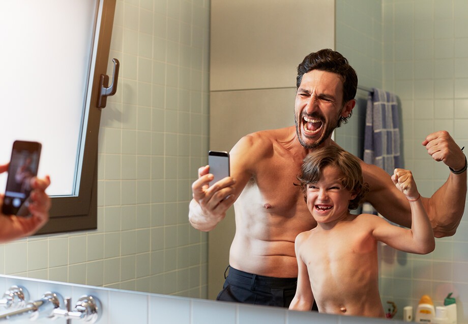Vater und Sohn stehen vor Spiegelwand im kleinen Badezimmer, zeigen die Muskeln und machen ein Selfie mit dem Handy 